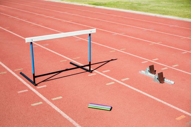 This image shows essential track and field equipment including a hurdle, relay baton, and starting block placed on a running track in a stadium. Ideal for use in sports training materials, athletic event promotions, fitness blogs, and educational content about track and field events.