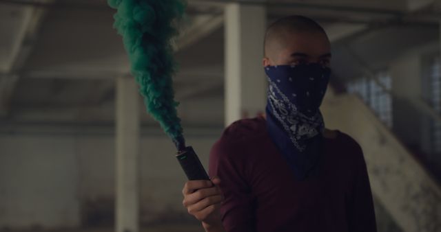 Young biracial man holds a green smoke flare, with copy space. Indoor setting adds a mysterious vibe as he covers his face with a bandana.