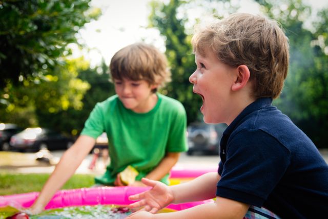 Two boys enjoying a sunny day playing outdoors near a kiddie pool. A child in a navy shirt laughs energetically while another boy in a green shirt is engaged in the background. Captures the essence of joyful, carefree childhood, ideal for use in advertisements, parenting blogs, summer activity promotions, or family lifestyle websites.