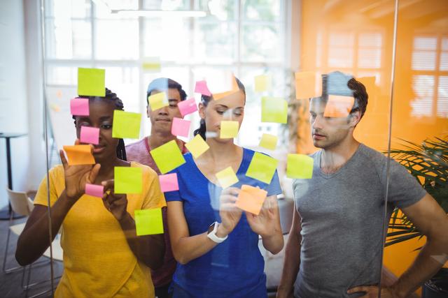 Diverse group of business professionals collaborating by sticking colorful sticky notes on glass wall in modern office. Ideal for illustrating concepts of teamwork, brainstorming sessions, project planning, and creative business strategies. Useful for business presentations, team-building workshops, and corporate training materials.