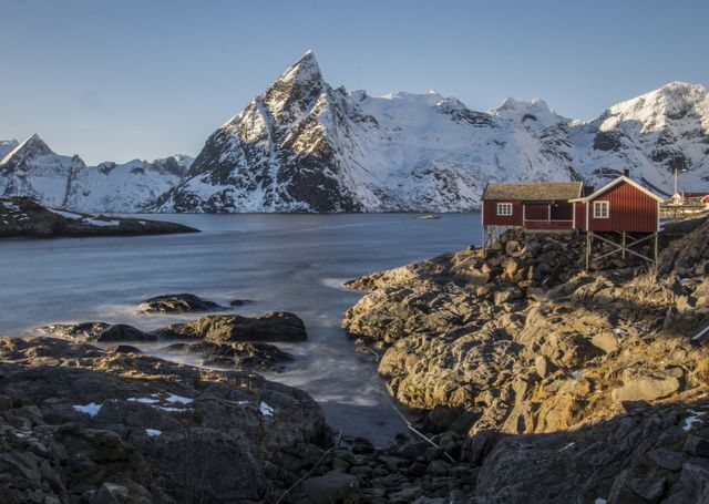 Red cabin perched on rocky shore overlooks serene river with significant snow-capped mountains rising in background. Ideal for illustrating remote cabin life, serene winter landscapes, nature excursions, and travel advertisements highlighting Scandinavia or cold winter retreats. Can be used in travel blogs, stock photography on outdoor living, or promotional materials for travel agencies.