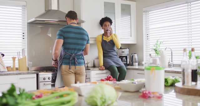 A cheerful couple is actively engaged in meal preparation in their modern kitchen. One person is cooking while the other is drinking water, showcasing a shared moment of domestic life. This image can be used for promoting healthy eating, relationship goals, home decor, and cooking content.