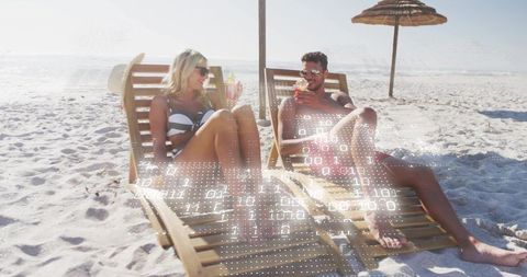 Couple relaxing on beach loungers with cocktails and digital data overlay