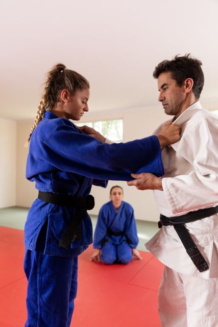 Caucasian male and female judokas wearing blue and white judogi, practicing judo on a mat during a training in a bright studio, while another female judoka is watching them in the background.