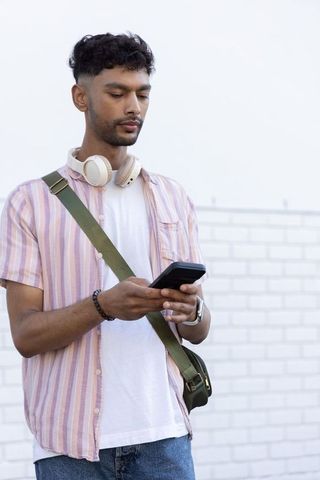 Asian mid-20s man standing outdoors using smartphone with headphones and messenger bag