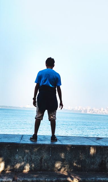 Person standing on seawall gazing at distant city skyline over calm ocean. Wearing casual clothing and facing away. Ideal for themes of solitude, contemplation, and urban scenery. Can be used in travel or lifestyle content.