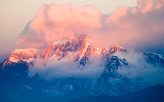Majestic mountain peak rising above clouds during sunset. Soft, glowing light casts warm hues on snow-capped mountains, creating a serene and breathtaking landscape. Ideal for nature-themed content, travel promotions, environmental campaigns, and backgrounds for websites or presentations featuring natural beauty.