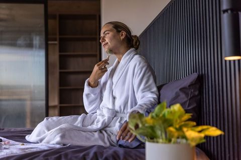 Woman wearing white bathrobe using facial roller on bed, relaxing morning skincare ritual