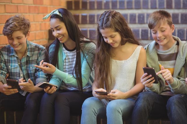 Group of smiling school friends sitting on staircase using mobile phone at school