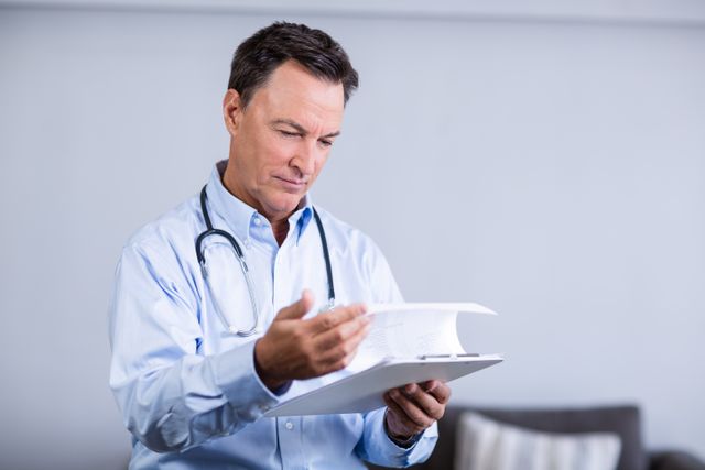 Male doctor reading reports in a clinic, wearing a stethoscope around his neck. Ideal for use in healthcare, medical, and professional settings. Can be used in articles, websites, and promotional materials related to healthcare, medical practices, and patient care.