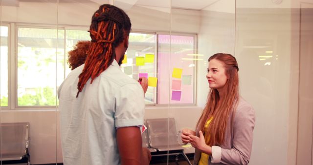 African American and Caucasian colleagues are engaged in a discussion in a bright office space, with copy space. They appear to be brainstorming ideas, as indicated by the sticky notes on the glass wall behind them.