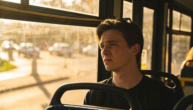 Young man gazing out bus window during golden hour commute, thoughtful urban traveler
