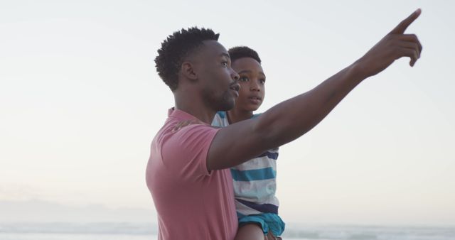 Man carrying young boy on beach while pointing towards something distant. Showcases family bonding, childhood experiences, and outdoor activities near ocean. Ideal for family-related advertisements, parenting articles, and lifestyle blogs.
