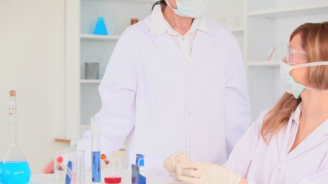 Two female scientists work collaboratively in a clinical laboratory, observing chemical reactions in test tubes and flasks. Their dedication and focus highlight the importance of teamwork and precision in scientific research. The setting underlines laboratory safety with both researchers wearing personal protective equipment. This visual can be useful for educational material, promoting careers in STEM fields, or emphasizing workplace safety standards.