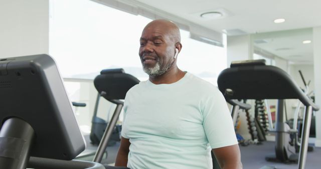 A senior man exercising on a treadmill at a modern gym, wearing a light blue shirt and white earbuds. Ideal for depicting fitness routines, promoting active and healthy lifestyle for seniors, gym advertisements, and health-related content.