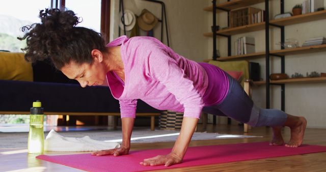 Woman engaging in a plank exercise on a pink yoga mat in a cozy living room. Ideal for content promoting home workouts, fitness tutorials, health blogs, and exercise routines.