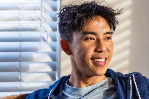 Young asian man smiling by window blinds warm daylight portrait casual blue hoodie