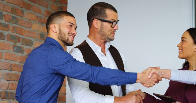 Three business people shaking hands and smiling in an industrial style office with a brick wall. This visual representation of collaboration and professional agreement can be used in articles or adverts about successful business partnerships, negotiations, teamwork, and corporate success stories.