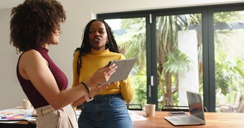 Female colleagues collaborating over tablet in bright modern workspace with garden view