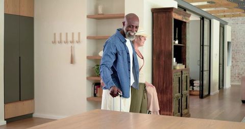 Mature diverse couple arriving at modern rustic home with rolling suitcase and brimmed hat