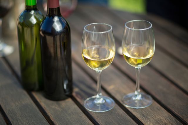 Close-up of two glasses of white wine and wine bottles on a wooden table. Ideal for use in articles or advertisements related to wine tasting, outdoor dining, summer parties, or elegant gatherings.