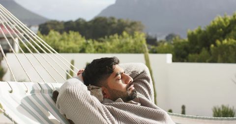 Indian man relaxing on hammock on sunny terrace wearing sweater, resting with mountain view