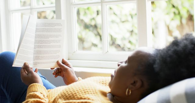 African American woman enjoying a quiet moment reading a book by a window. Readers, leisure blogs, relaxation articles, and promotional material on hobbies or home lifestyle can use this.