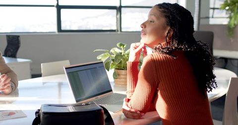 Diverse female colleague leaning and collaborating at sunlit modern office table with laptop