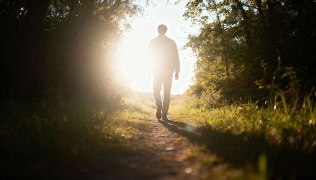 Solitary man walking through sunlit forest trail casting long shadow and warm sun flare