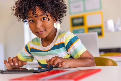Curly-haired african american boy using tablet at classroom desk, learning and focusing