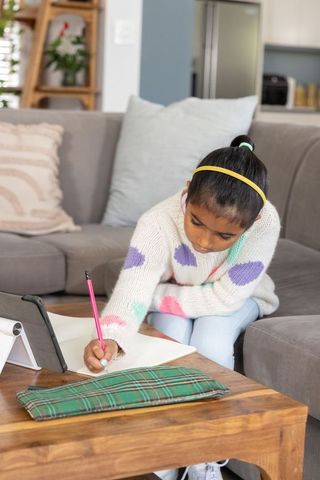Indian girl writing homework with tablet on gray sofa, studying at home in cozy living room