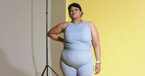 Plus-size model posing in blue ribbed activewear with black cap and smartwatch on yellow backdrop
