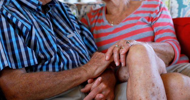An elderly Caucasian couple sits closely, holding hands in a gesture of comfort and long-standing companionship. Their clasped hands and matching rings speak volumes about their enduring relationship and mutual support through the years.