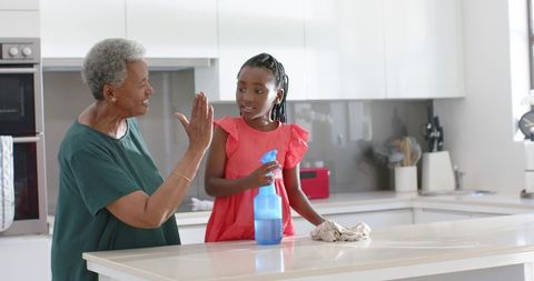 African american grandmother and granddaughter high-fiving while cleaning sunlit kitchen