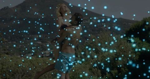 Couple embracing on sunlit hillside, man lifting woman in turquoise trunks, romantic glow