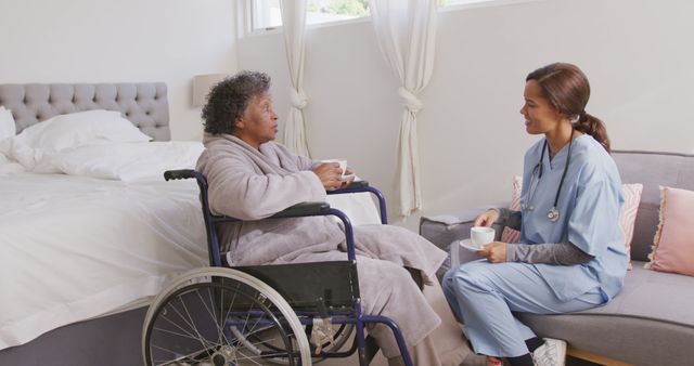 Senior african american woman sitting in wheelchair and female doctor drinking coffee and talking. Medicine, healthcare, disability and lifestyle.