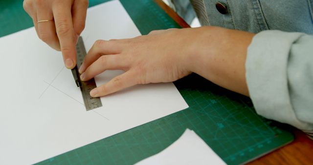 Close-up showing person using craft knife and ruler to cut along lines on white paper, placed on green cutting mat. Useful for DIY, Crafts, precision cutting, instructional guides, art projects, stationery, accurate measurements, hobbies, and creative activities
