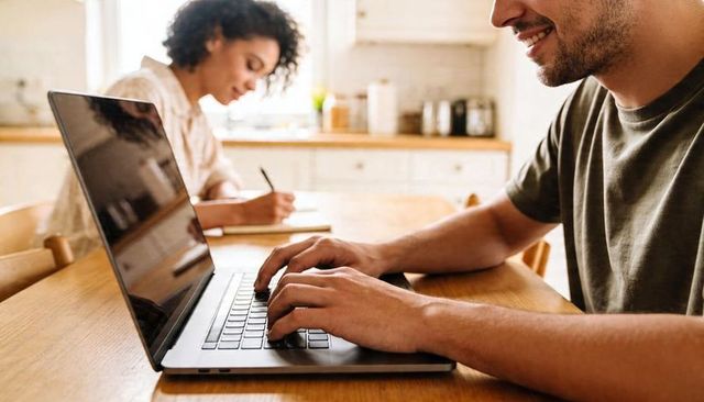 Man typing on laptop while woman writing at sunlit kitchen table for remote work and study