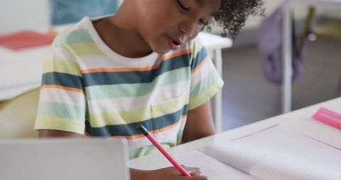 Primary student writing in notebook while using laptop and pink pencil in classroom