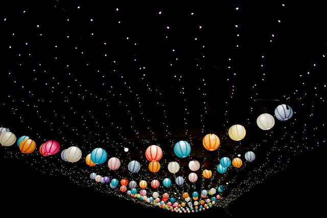 Colorful paper lanterns hanging under fairy lights, creating a vibrant and festive atmosphere in the night sky. Suitable for use in articles or promotions related to festivals, outdoor parties, celebrations, decorative lighting, or creative inspirations for event planning.