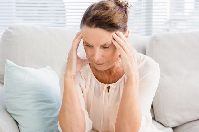 Mature woman sitting on sofa with head in hands, appearing stressed or thoughtful. Ideal for use in articles about mental health, stress management, aging, or personal struggles. Can be used in blogs, websites, or advertisements related to wellness, healthcare, or lifestyle.
