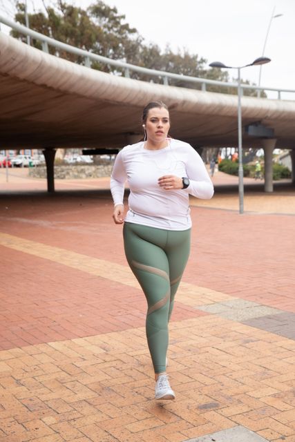 A curvy Caucasian woman with long dark hair wearing sports clothes is running with earphones on in an urban pedestrian area. This image is perfect for use in fitness-related content, promoting active lifestyles, and showcasing diversity in body types in sports and exercise. It can also be used in campaigns emphasizing outdoor activities and urban fitness routines.