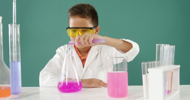 A young boy wearing protective glasses and a white lab coat pours a pink liquid into a beaker while conducting a science experiment. The scene includes various laboratory glassware filled with vibrant colored liquids. Ideal for educational materials, promoting STEM education, classroom posters, or science-related websites.