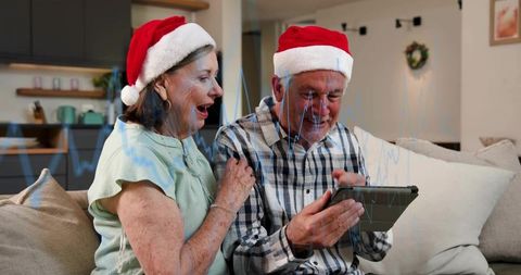 Senior couple laughing and sharing tablet in cozy living room wearing santa hats