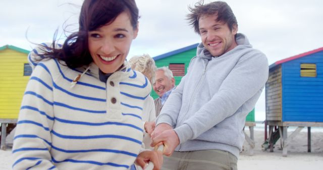 A joyful young Caucasian couple is holding hands on a beach with colorful beach huts in the background, with copy space. Their smiles and playful demeanor suggest a moment of happiness and leisure during a beach outing.