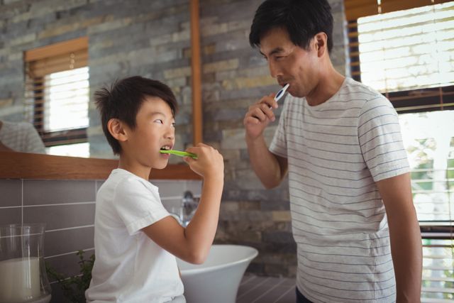 Father and son are standing in a modern bathroom, brushing their teeth together. The intimacy and bonding during daily hygiene routines emphasize strong family relationships. This could be used in advertisements for dental care products, parenting articles, or health and wellness promotions.