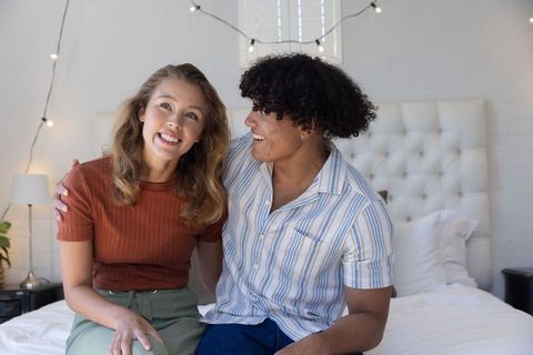 Diverse young couple sitting on bed, smiling and embracing under string lights, cozy bedroom