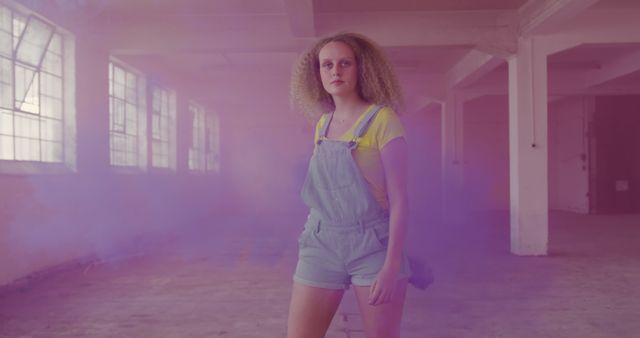 Confident woman with curly hair wearing casual clothing stands in a purple haze inside an abandoned warehouse. Ideal for themes of resilience, modern fashion, creative concepts, and artistic expressions. Great for use in fashion editorials, creative projects, and urban-themed designs.