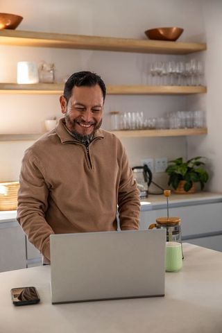 Mature asian man working from home at kitchen island with laptop, french press and mug