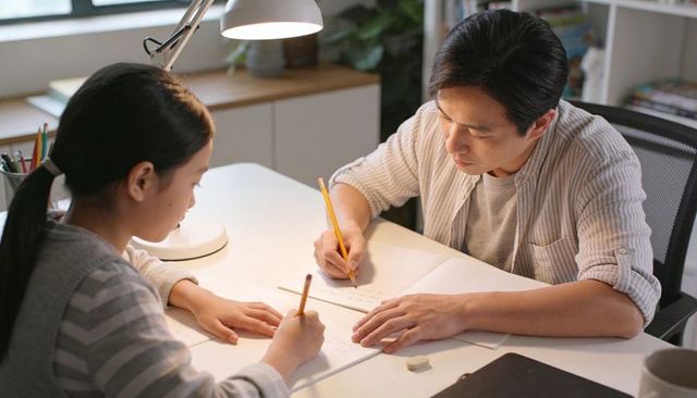 Asian father helping daughter with homework at home desk under lamp studying together